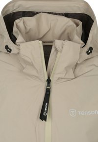 Beige waterdichte jas met een hoge kraag, ritssluiting aan de voorkant, verstelbare toggles en een logo-detail. Gladde, lichte stof textuur.