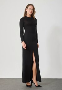 Μαύρο φόρεμα με μακριά μανίκια και ψηλή σχισμή, κατασκευασμένο από απαλό ύφασμα, με εφαρμοστή γραμμή και ελάχιστες λεπτομέρειες.