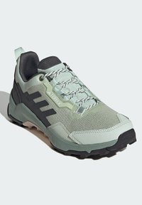 Zapatilla deportiva verde claro con parte superior de malla, acentos negros y suela de goma. Presenta áreas texturizadas y cordones para un ajuste ceñido.