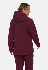 Bordeaux utomhusjacka med huva, dragkedjsidofickor och en logotyp på den övre ryggen. Slät textur och åtsittande design för värme.