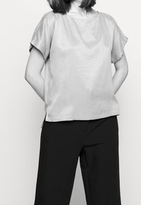 Donna che indossa una blusa larga a maniche corte, lasciata fuori dai pantaloni neri a gamba larga, con le mani dietro la schiena, su uno sfondo neutro.
