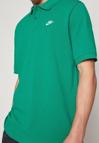Man som bär en grön Nike polo skjorta med vitt logo på vänster bröst, står med händerna i fickorna mot en enfärgad bakgrund.