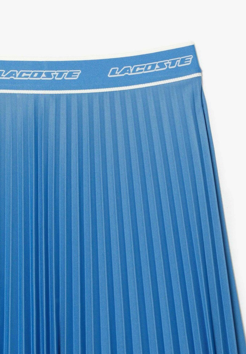 Jupe plissée bleue en tissu texturé, avec une bande logo blanche à la taille et de distincts plis verticaux sur l'ensemble du design.