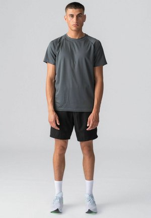 Unge mand står iført en mørkegrå atletik t-shirt, sorte shorts, hvide sokker og hvide løbesko mod en ensartet lys baggrund.