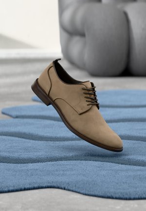 Beżowe, sznurowane eleganckie buty unoszące się nad teksturowanym niebieskim dywanem, w tle rozmyte, szare, tapicerowane krzesło.