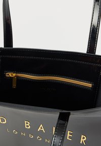 Črna torbica iz lakiranega usnja z zlatim žepom z zadrgo in vtisnjenim logotipom. Notranjost z gladkim otipom je odprta in prikazuje organizacijske funkcije.