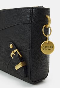 Coin de sac en cuir noir texturé avec détail en boucle dorée et étiquette ronde en or de la marque Guess gravée "GUESS EST 1981" attachée par un anneau doré.