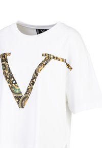Weißes Baumwoll-T-Shirt mit einem großen schwarzen und goldenen Paisleymuster "V" auf der Vorderseite. Kurze Ärmel und ein lässiger Schnitt.