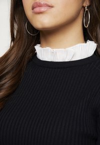 Svart ribbad tröja med vit volangkrage. Modellen bär silverfärgade hoopörhängen. Mjuk textur och lagerdesign synlig.