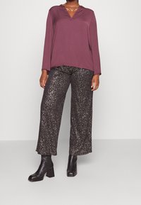 Maglietta a maniche lunghe color marrone con scollatura a V, abbinata a pantaloni neri larghi con glitter. Stivali neri alla caviglia con tacco spesso.