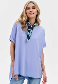 Jasnofioletowa, oversize'owa bluzka z podwiniętymi rękawami i okrągłym dekoltem, zestawiona ze wzorzystym czarno-niebieskim szalem. Gładka faktura, luźny krój.