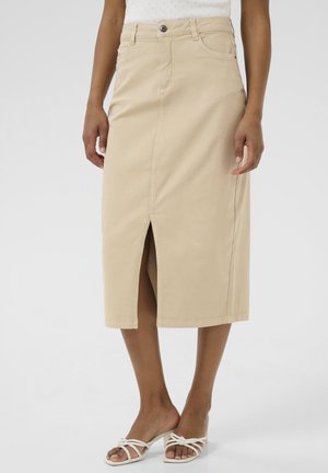 Kvinde iført en beige midi-nederdel med en frontslids, en hvid top og hvide sandaletter med hæl, stående mod en ensfarvet baggrund.