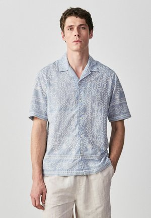 Junger Mann, der ein hellblaues, kurzärmeliges Hemd mit weißer Blumenstickerei und eine hellbeige Hose trägt, steht vor einem einfarbigen Hintergrund.