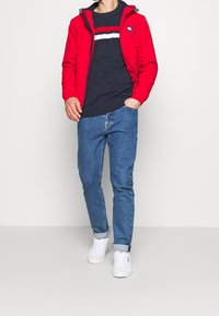 Veste zippée rouge avec capuche ; pull bleu marine avec des rayures blanches et rouges ; jean bleu ; baskets blanches avec les manches retroussées ; tenue moderne décontractée.