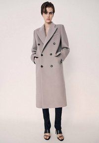 Lang dobbeltknappet frakke i lys grå uldblanding med store revers, sorte knapper og sidelommer, kombineret med smalle sorte bukser og hæle.