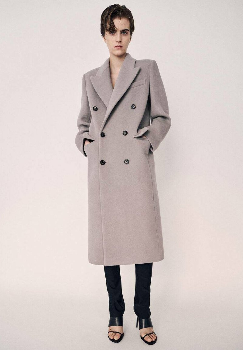 Lang dobbeltknappet frakke i lys grå uldblanding med store revers, sorte knapper og sidelommer, kombineret med smalle sorte bukser og hæle.
