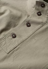 Tejido de algodón beige con una superficie texturizada, que presenta una tapeta con botones marrones y un cuello puntiagudo con costura detallada.