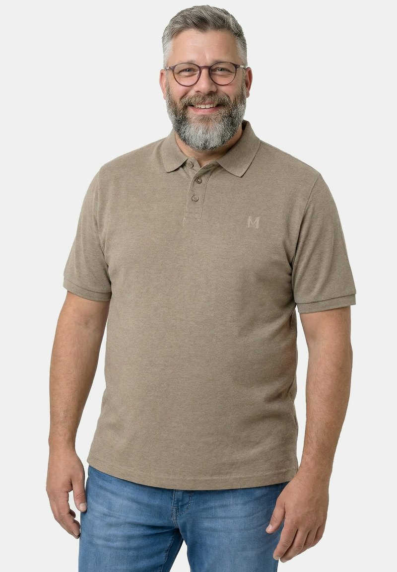 Uomo di mezza età sorridente con barba grigia e occhiali, che indossa una polo beige e jeans blu, in piedi davanti a uno sfondo bianco uniforme.