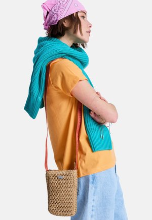 Femme en t-shirt orange et jean bleu portant un bandana rose et un pull bleu sarcelle, avec un sac bandoulière tressé à sangle rouge.