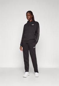 Schwarzer Kapuzensweatshirt und passende Jogginghose, beide mit einem auffälligen Logo. Weicher Stoff mit weißen seitlichen Akzenten und einer Kängurutasche.