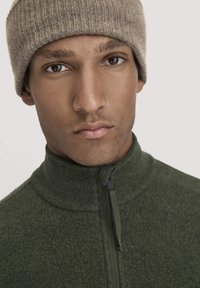 Grüne Fleecejacke mit hohem Kragen und Reißverschluss, kombiniert mit einer beigen Rippstrickmütze. Die Textur wirkt weich und warm.