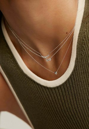 Collares de plata en capas con diseños circulares y adornados, cayendo sobre una blusa texturizada verde oliva con un escote crema.