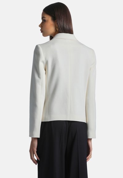 Femme aux cheveux lisses et foncés, portant un blazer blanc et un pantalon noir, vue de dos, tournée vers la gauche sur un fond uni.