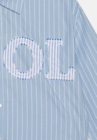 Camicia a righe blu chiaro con bottoni bianchi, caratterizzata da grandi lettere ricamate "O" e "L" con un motivo a quadri blu e bianchi.