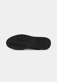 Semelle de chaussure en caoutchouc noir avec grip texturé et logo "Tommy Jeans" embossé près de la zone des orteils sur fond blanc.