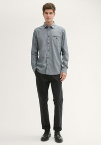 Hemd mit Knopfleiste aus grauem Stoff, lange Ärmel, Brusttasche. Getragen mit schwarzen Hosen und schwarzen Schuhen. Einfaches Design, keine sichtbaren Muster.