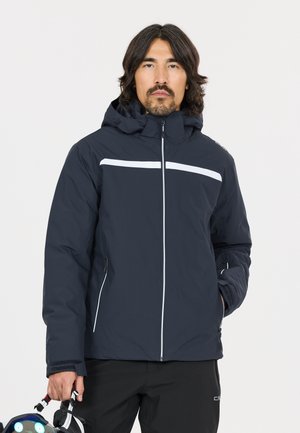Mand iført en marineblå jakke med hvide reflekser, der holder skibriller og hjelm i den ene hånd mod en ensartet lys baggrund.