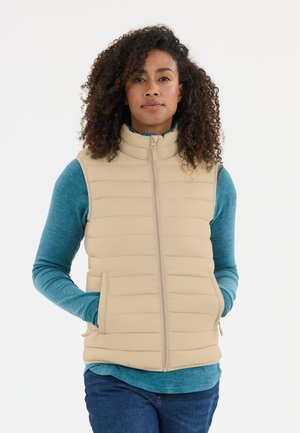 Vrouw met krullend haar die een beige doorgestikte vest draagt over een blauw overhemd met lange mouwen en een blauwe spijkerbroek, handen in de vestzakken.