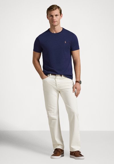 T-shirt blu navy a maniche corte con un piccolo logo, abbinata a pantaloni beige chiaro e sneaker marroni, creando un look casual.