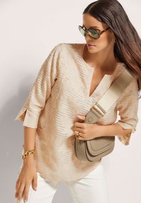 Beige strukturierter Oberteil mit V-Ausschnitt und Dreiviertelärmel, kombiniert mit einer kleinen beigefarbenen Tasche. Akzente setzen ein goldener Kettenarmreif und Ringe.