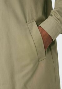 Veste légère et verte avec une poche latérale et une texture lisse. La manche présente un détail de poignet cousu, soulignant un design pratique.