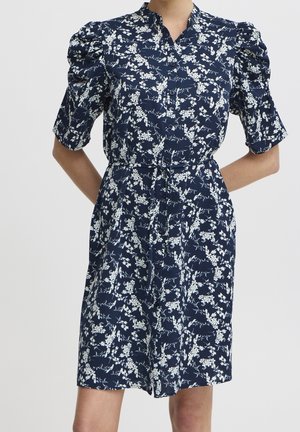 Woman wearing navy blue button-up dress with white floral pattern, puffed short sleeves, and waist tie, standing with hands behind back.