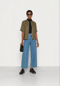Camisa verde de mangas curtas sobre um top preto, combinada com jeans azuis de perna larga de cintura alta e sapatos pretos robustos. Inclui uma bolsa preta e óculos de sol.
