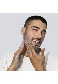 Produkt do oczyszczania twarzy pokazywany podczas aplikacji przez osobę. Jasnoszara pianka na twarzy, krótkie ciemne włosy i ubrana w kremową koszulę.