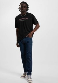 Fekete, rövid ujjú póló rózsaszín "ROCAWEAR" logóval, kék farmernadrággal és szürke sportcipővel, fehér díszítéssel és fűzővel párosítva.
