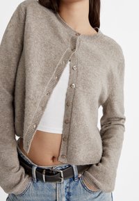 Cardigan in misto lana beige con scollatura rotonda, chiusura a bouton, polsini a costine e leggero sovrapporsi nella parte anteriore. Indossato sopra un top bianco.