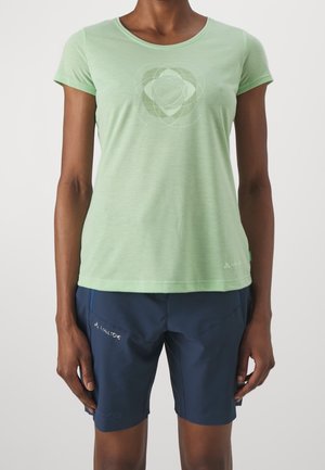 Osoba ubrana w jasnozieloną koszulkę z krótkim rękawem z delikatnym geometrycznym logo oraz granatowe szorty z bocznymi kieszeniami.