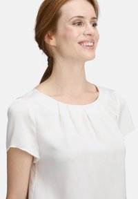 Weiße kurzärmlige Bluse mit Rundhalsausschnitt und sanften Falten an der Vorderseite, aus einem leichten Stoff mit glatter Textur.