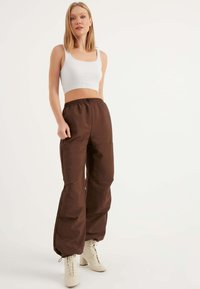 Braune, lockere, leichte Hose mit elastischem Bund. Kombiniert mit einem weißen, gerippten Croptop und beigen geschnürten Absatzstiefeln.