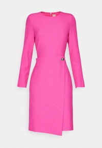 Robe rose vif longueur genou avec manches longues, encolure ronde et rabat asymétrique à l’avant fixé par un seul bouton à la taille.