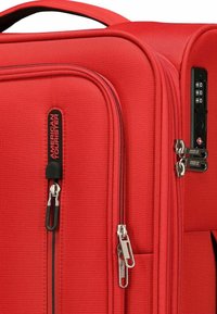 Valise rigide rouge avec une finition texturée, double zip, serrure à combinaison intégrée et une poignée de transport supérieure. Présente une étiquette de marque.