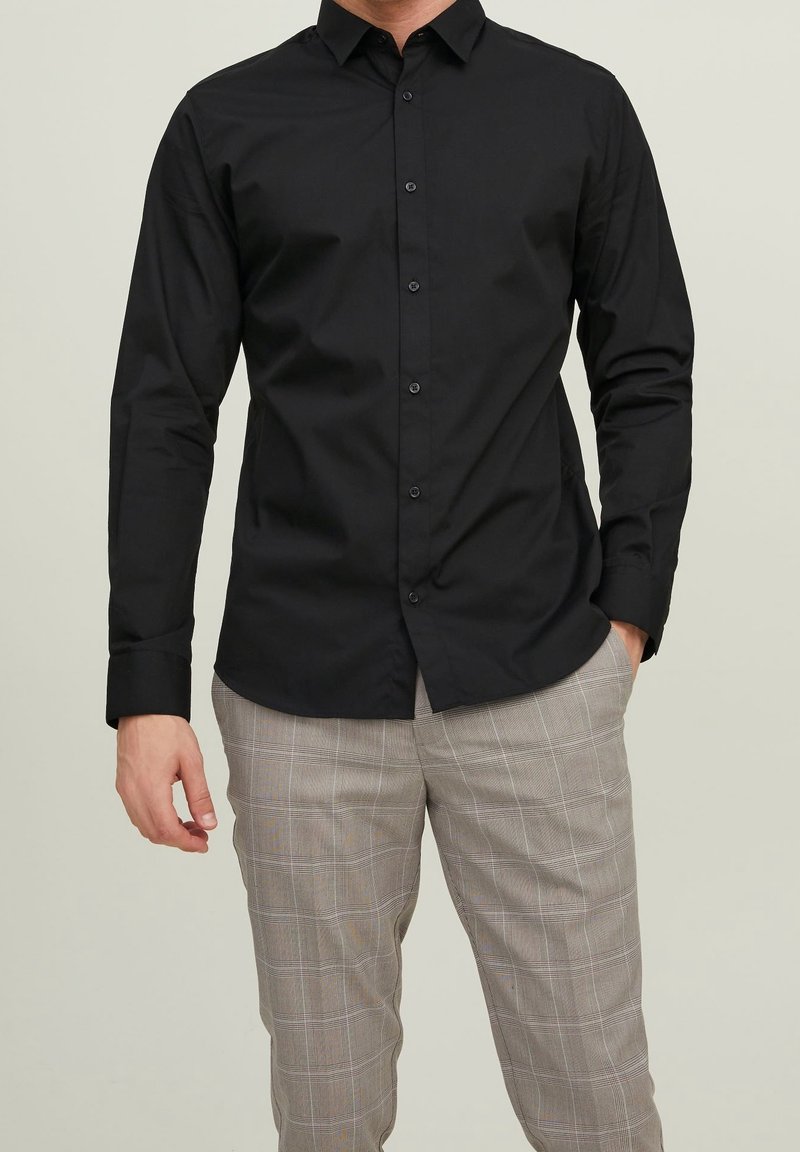 Camicia nera a maniche lunghe; design con bottoni; realizzata in tessuto liscio; presenta un colletto classico ed è abbinata a pantaloni grigi a quadri.