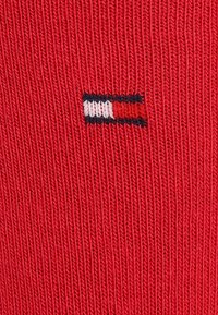 Tissu tricoté rouge présentant un petit logo brodé avec des rayures horizontales bleues, blanches et rouges et un détail en contour noir.