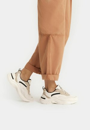 Person wearing beige and white chunky sneakers with black accents, paired with rolled-up light brown pants against a plain background.