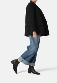 Blazer negro, jeans azules holgados con puños doblados y botas negras de tobillo con un tacón bajo. La tela suave y el corte entallado acentúan la figura.