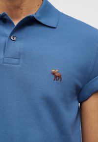 Μπλε πόλο πουκάμισο με μανίκια κοντά γυρισμένα και μικρό κεντημένο καφέ ελάφι στο αριστερό στήθος, φορεμένο από ένα άτομο.